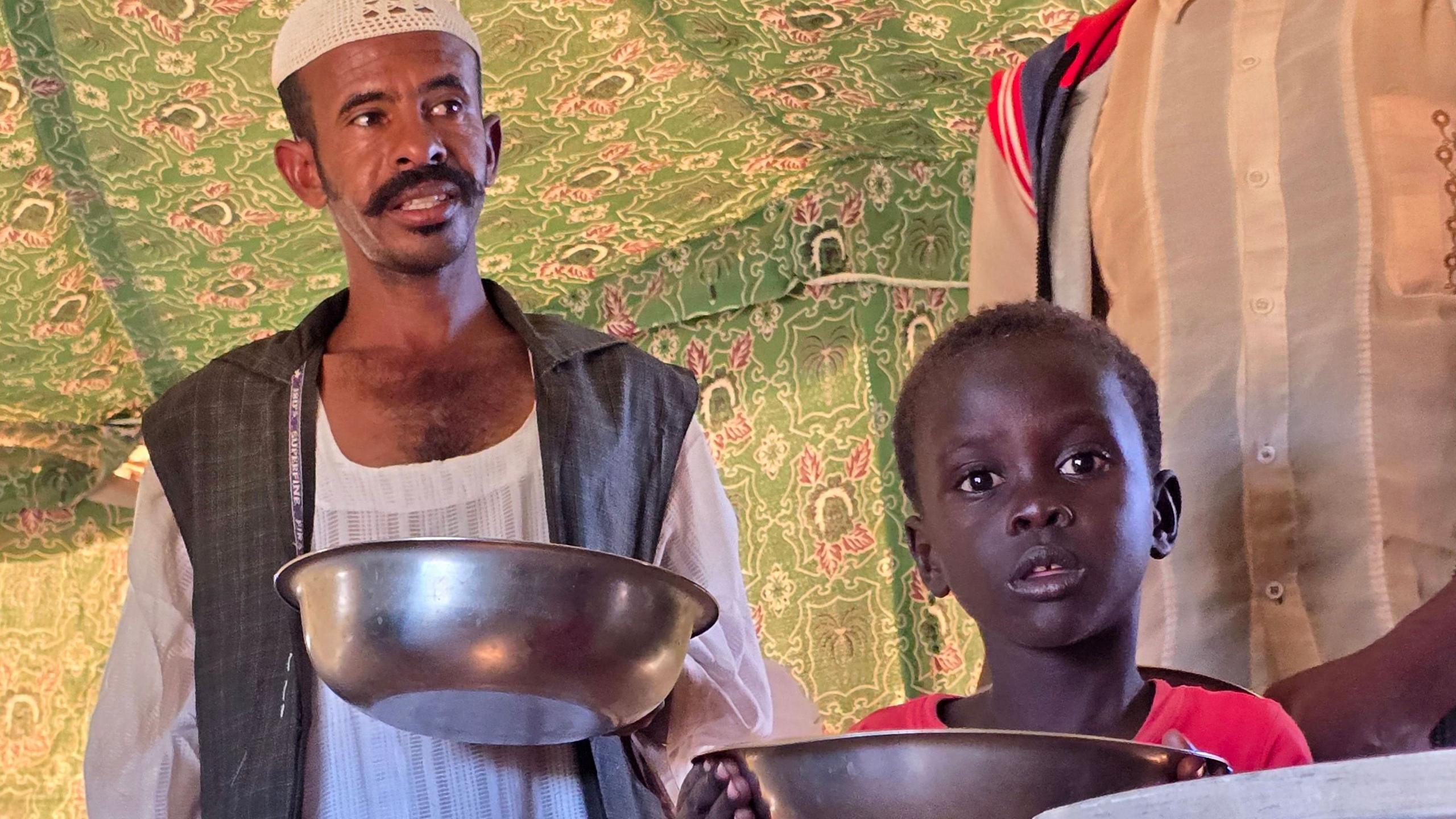 A man in a white robe holds a bowl as he waits for aid alongside a young boy in a red T‑shirt.