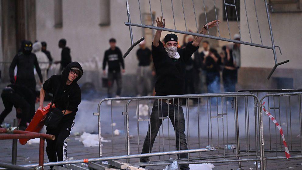 Man in black hoodie throwing a barrier up over another while a man stands to his right and holds a red object in Milan