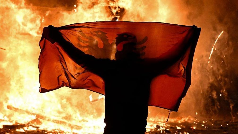 Protester holding Albanian flag in front of a burning kiosk in Tirana
